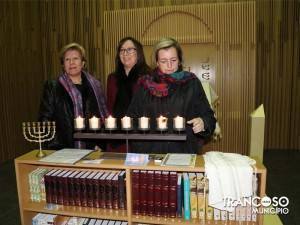 Três mulheres junto a uma menorá acesa e livros religiosos numa sala com painéis dourados