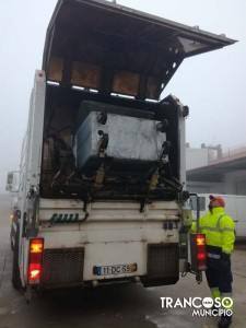 Camião de recolha de resíduos com comporta traseira elevada, contentor de carga visível, operário em colete fluorescente ao lado.