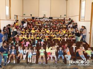 Centenas de crianças e adultos sentados em anfiteatro escuro durante apresentação teatral, muitas crianças com camisolas amarelas.