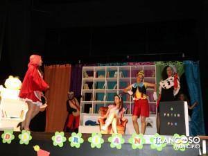 Cena de peça teatral infantil com atores vestidos como personagens de contos, incluindo uma rapariga de capa vermelha, em cenário com estantes de livros coloridas.