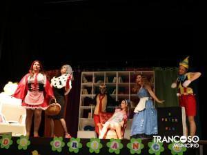 Cinco atores em palco representando cena de teatro infantil, com cenário com estantes de livros e decorações coloridas ao fundo.