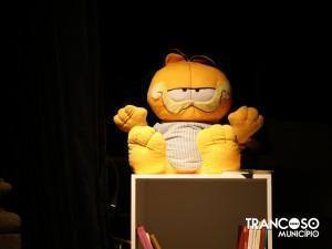 Boneco de Garfield cor-de-laranja com óculos escuros e patas amarelas sentado sobre uma estrutura castanha, em cenário de teatro com iluminação de palco.