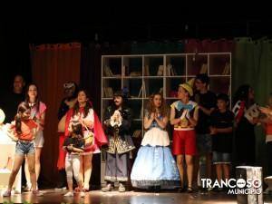 Grupo de crianças e adolescentes em cena teatral, usando várias fantasias coloridas, num palco com estante de livros ao fundo.