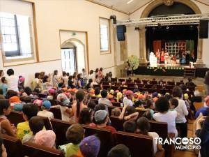 Crianças sentadas em bancos de madeira na plateia de um templo, viradas para um palco iluminado onde decorre uma representação teatral.