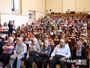 Auditório cheio com crianças, adultos e idosos sentados em poltronas castanhas, assistindo a evento teatral.