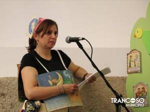 Mulher com fita decorativa na cabeça lê livro ao microfone, durante apresentação infantil com painéis temáticos ao fundo.