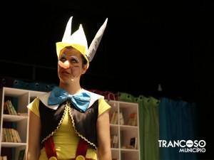 Criança caracterizada como personagem de conto de fadas, com coroa branca e amarela, veste amarela e laço azul, em palco com estantes de livros ao fundo.
