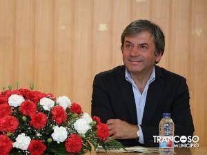 Homem de meia-idade em blazer preto e camisa azul sentado perante arranjo de cravos vermelhos e brancos, com garrafa de água à sua frente.