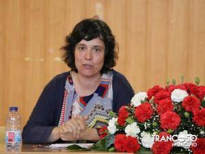 Mulher de meia-idade a falar em pódio, com arranjo floral de cravos vermelhos e brancos à sua frente e garrafa de água à esquerda.