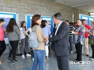 Homem de fato em conversação com mulher grávida durante cerimónia de abertura de exposição, rodeados por visitantes junto a painéis informativos azuis.