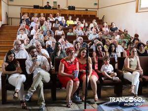 Auditório cheio com dezenas de pessoas sentadas em filas de bancos de madeira durante apresentação de livro