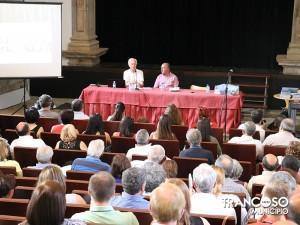 Dois oradores sentados atrás de mesa vermelha num auditório, com público a assistir de costas para a câmara.