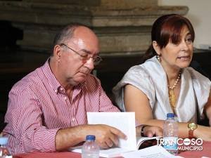 Dois oradores sentados em painel de apresentação, homem de óculos em camisa rosa e mulher de blusa branca, junto a microfones e garrafas de água.