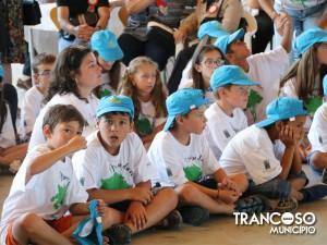 Grupo de crianças sentadas no chão com camisetas brancas e bonés azuis, num evento comunitário