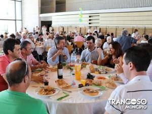 Grupo de pessoas a comer e conversar em mesas redondas durante evento em pavilhão desportivo.