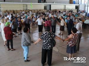 Grupo diversificado de pessoas idosas dança em círculo num pavilhão desportivo com mãos dadas.