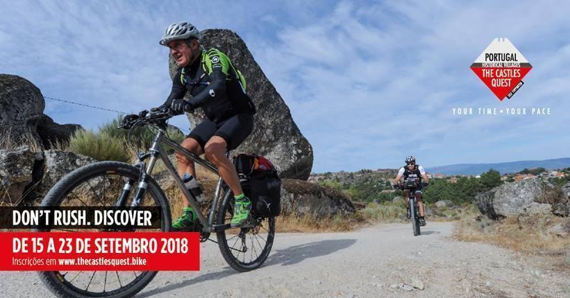 Ciclista de capacete e mochila verde em estrada rural portuguesa, com rochas graníticas e paisagem montanhosa ao fundo.
