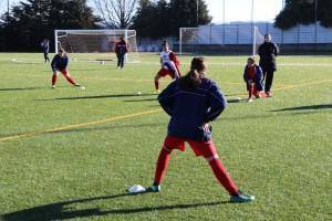 Rapariga em posição de defesa durante treino de futebol com outras crianças num campo com balizas e redes.