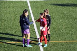 Duas raparigas de equipas diferentes (uma em uniforme roxo, outra em vermelho) encontram-se no centro do campo de futebol antes da partida.