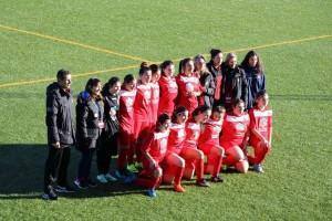 Equipa de futebol feminino em uniforme vermelho alinhada no campo com equipa técnica ao lado.