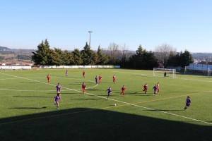 Duas equipas de jovens em jogo de futebol num campo desportivo municipal, uma em vermelho e outra em roxo.