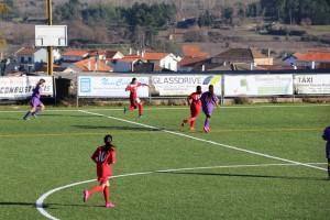 Jogadores em campo durante partida de futebol, rapazes em equipamentos vermelho e roxo, vila com casarões ao fundo