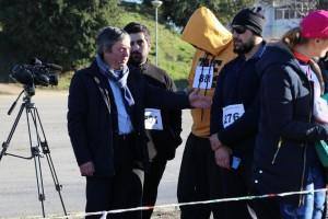 Homens em casacos pretos e participantes com dorsais conversam junto a câmara de televisão profissional durante evento desportivo.