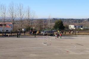 Crianças e jovens a jogar futebol num campo descoberto com balizas de treino, rodeados de espectadores e transporte escolar ao fundo.