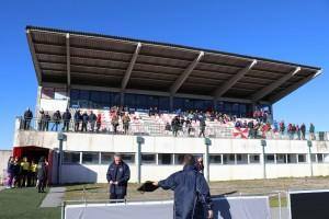 Estádio com bancada coberta abrigando espectadores, campo com atletas em ação e estrutura moderna de cobertura solar.