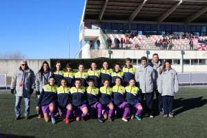 Equipa de futebol feminino em uniforme amarelo, azul e roxo, junto com treinadores e staff, perfilada no relvado de um estádio com bancadas ao fundo.
