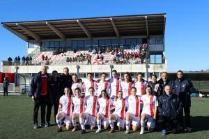 Equipa de futebol feminino em uniforme branco e vermelho com membros da comissão técnica, posicionados em frente a estádio com adeptos nas bancadas.