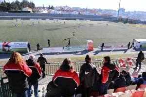 Adeptos em bancada com casacos vermelhos e pretos a observar partida de futebol em campo com cidade ao fundo
