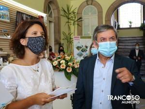 Mulher de máscara preta com padrão e homem de máscara azul e blazer em edifício histórico com arcos e plantas.
