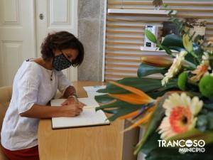 Mulher de máscara assinando documento numa mesa de madeira, com flores coloridas em primeiro plano e divisória de madeira ao fundo.