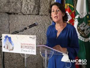 Mulher de blusa azul a falar ao microfone, com podium de acrílico, bandeiras municipais ao fundo e logótipo de Trancoso visível.
