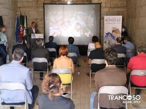 Apresentação em auditório com palrador, plateia sentada e projeção de imagens em ecrã. Bandeiras ao fundo e cartaz sobre Rupestre visível.