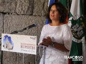 Mulher em traje branco a falar ao microfone com cartaz de necrópole em fundo, bandeiras de Trancoso atrás.
