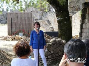 Mulher em blazer azul ao microfone, com público de costas, junto a ruínas medievais e muralha de pedra.