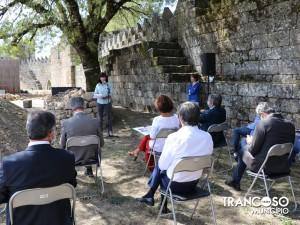 Grupo de pessoas sentadas em cadeiras numa zona arqueológica junto a muralhas de pedra, com duas mulheres em pé a dirigir-se à audiência.