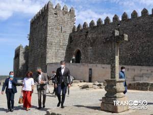 Grupo de pessoas em traje formal caminha junto à muralha medieval do Castelo de Trancoso, com uma estrutura moderna junto à base da fortificação.