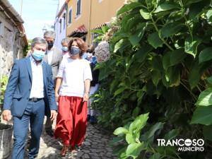 Secretária de Estado do Turismo caminha em rua medieval de Trancoso acompanhada por comitiva oficial, com edifícios históricos e vegetação verdejante.