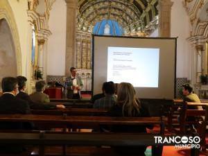 Apresentador em pé junto a ecrã de projeção numa nave de igreja historicamente decorada, com assistentes sentados em bancos de madeira a ouvir.