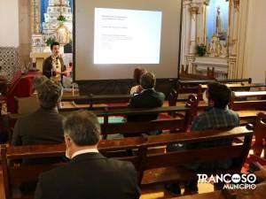 Apresentador gesticula enquanto fala junto a ecrã de projeção numa cappela, perante vários assistentes sentados em bancos de madeira. Ao fundo, imagens religiosas e altar.