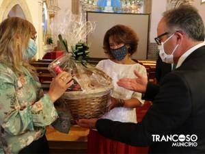 Duas mulheres e um homem de máscara, num interior de igreja, entregam cesto com produtos embrulhados a uma mulher.