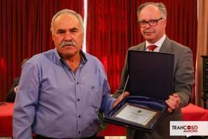 Dois homens numa cerimónia: um de camisa azul à esquerda e outro de fato cinzento segurando uma caixa azul-escuro aberta com certificado.
